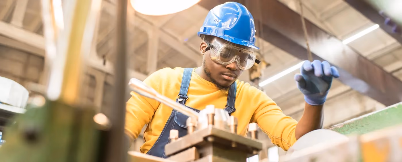 Worker in blue hardhat and goggles operating machinery in an industrial workshop.
