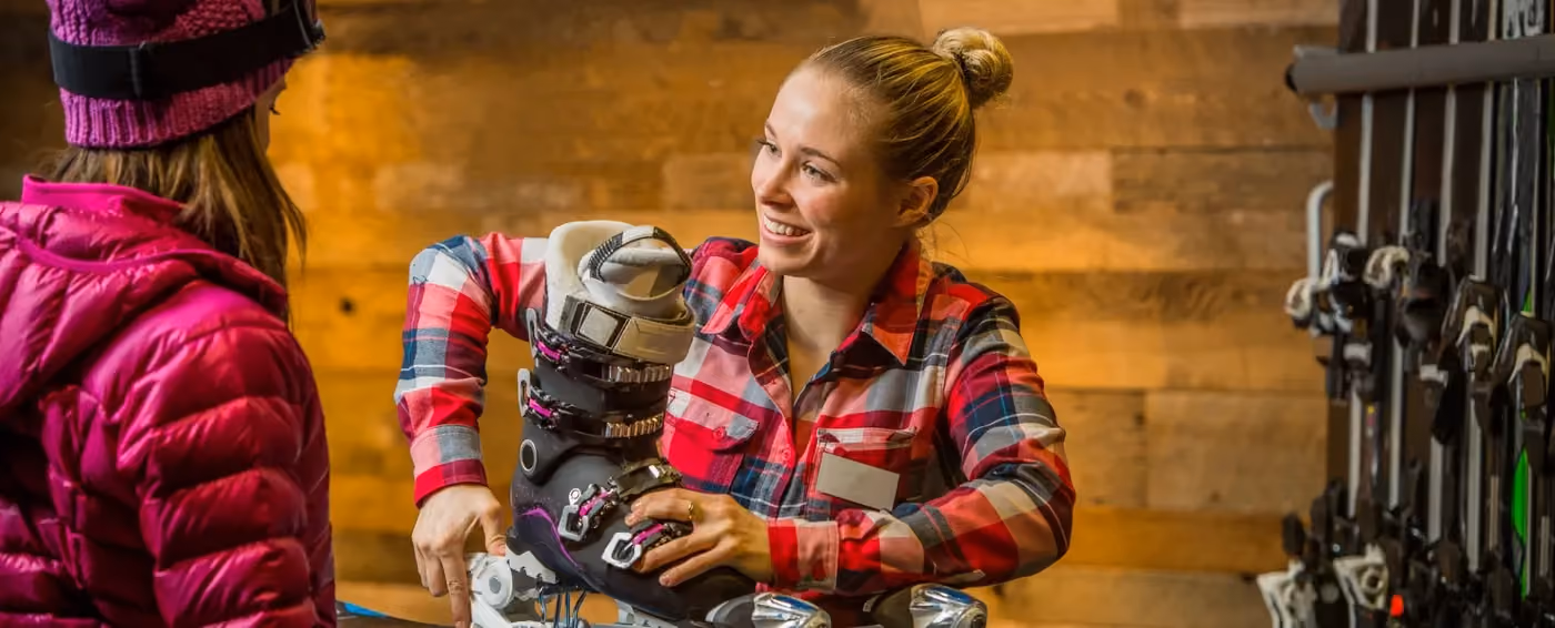 Smiling woman in plaid fitting a ski boot onto a customer's foot near ski equipment.