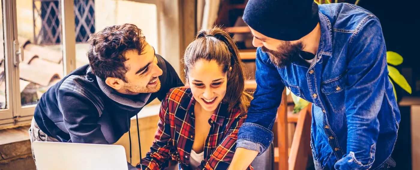 Three young coworkers smiling and collaborating on a laptop in a bright office space.