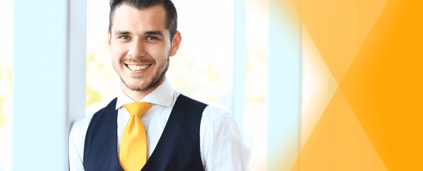 Smiling man in white shirt, navy vest, and yellow tie standing by a window with orange graphics.