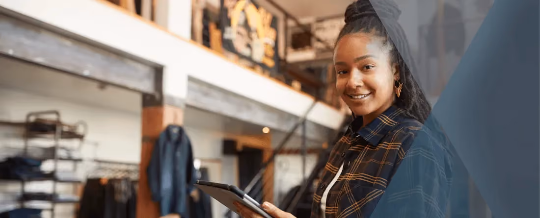 Smiling woman holding a tablet in a casual retail or workspace setting with shelves and jackets behind her.