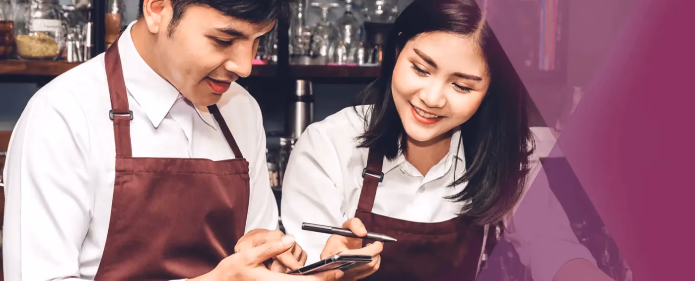 Two cafe workers in white shirts and brown aprons looking at a phone and smiling.