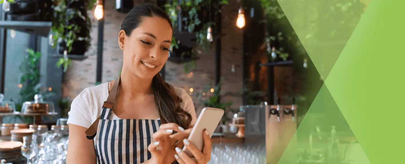Smiling woman in striped apron using smartphone in a café with plants and glassware in background.