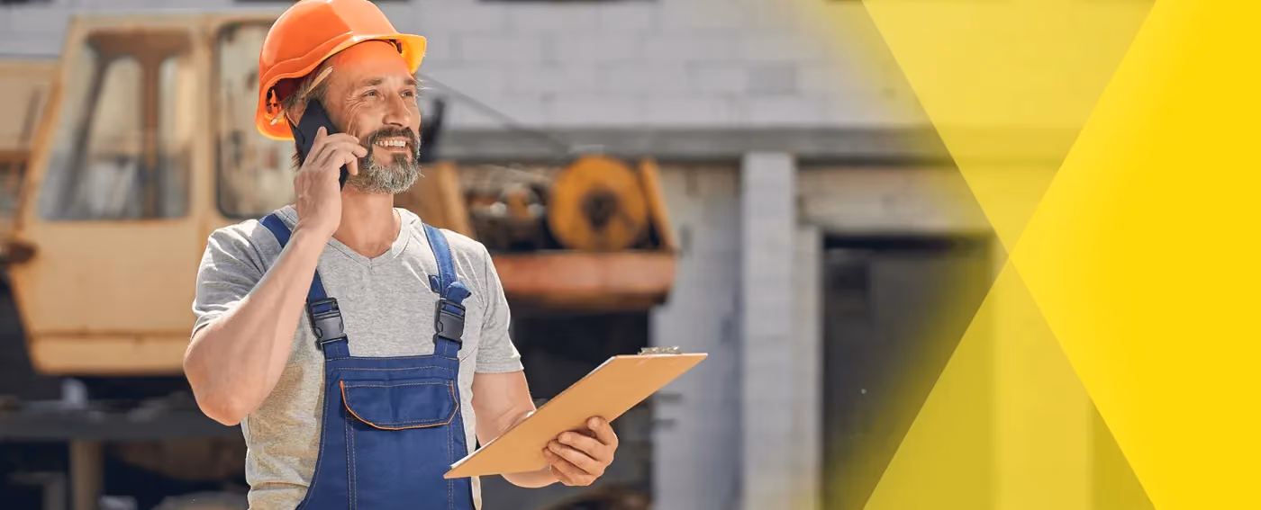 Construction worker in orange helmet and overalls talking on phone with clipboard in hand at worksite.