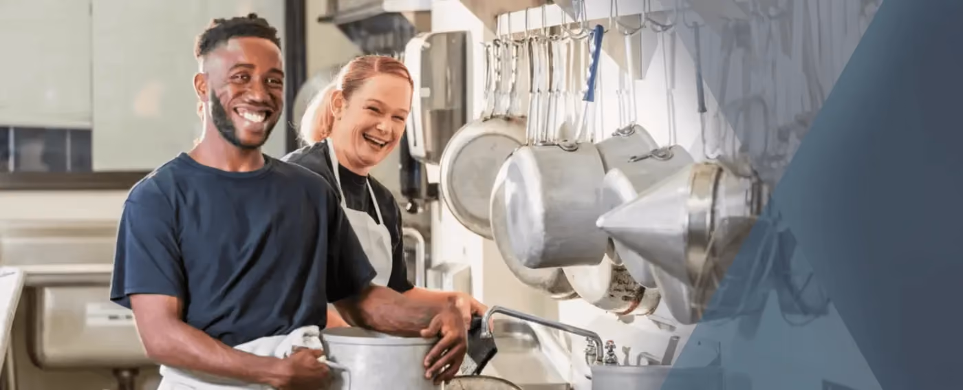 Two cheerful kitchen staff washing dishes with hanging pots and pans in the background.