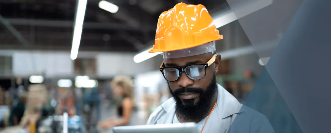 Bearded man wearing orange hard hat and glasses working on tablet in industrial warehouse.
