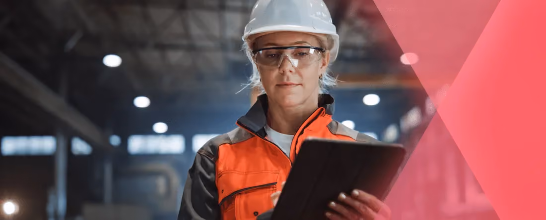 Woman in safety gear and hard hat using a tablet in an industrial warehouse setting.