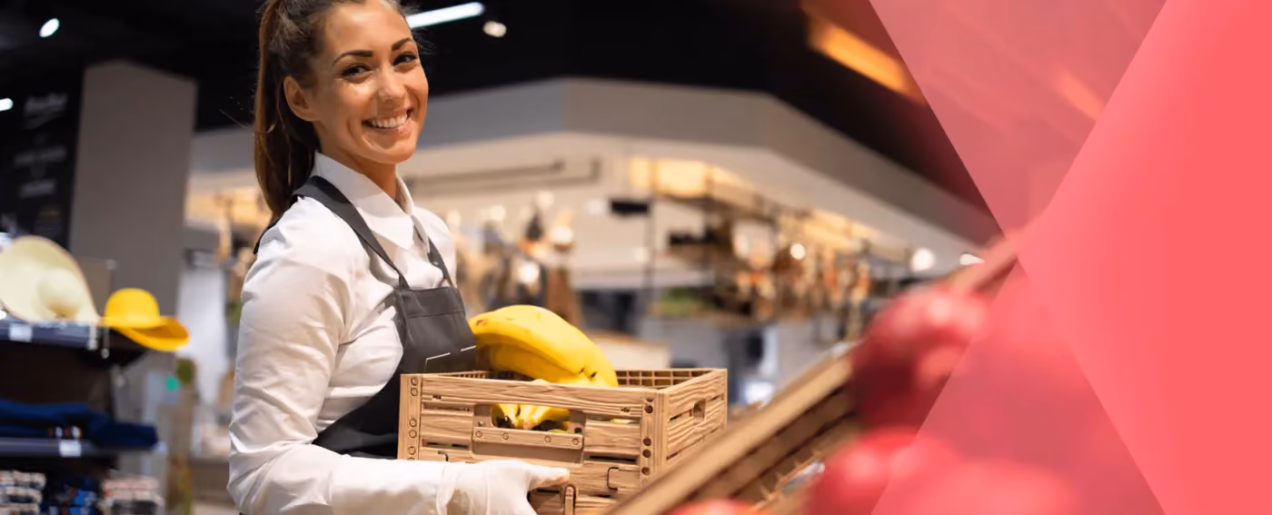 Smiling woman in apron holding crate with bananas inside a grocery store.