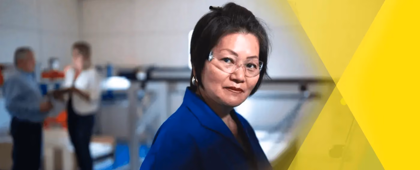 Woman in blue work uniform and safety glasses standing in an industrial or manufacturing setting.
