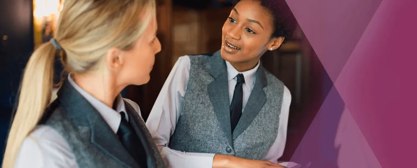 Two female hospitality workers in gray vests and ties engaged in conversation indoors.