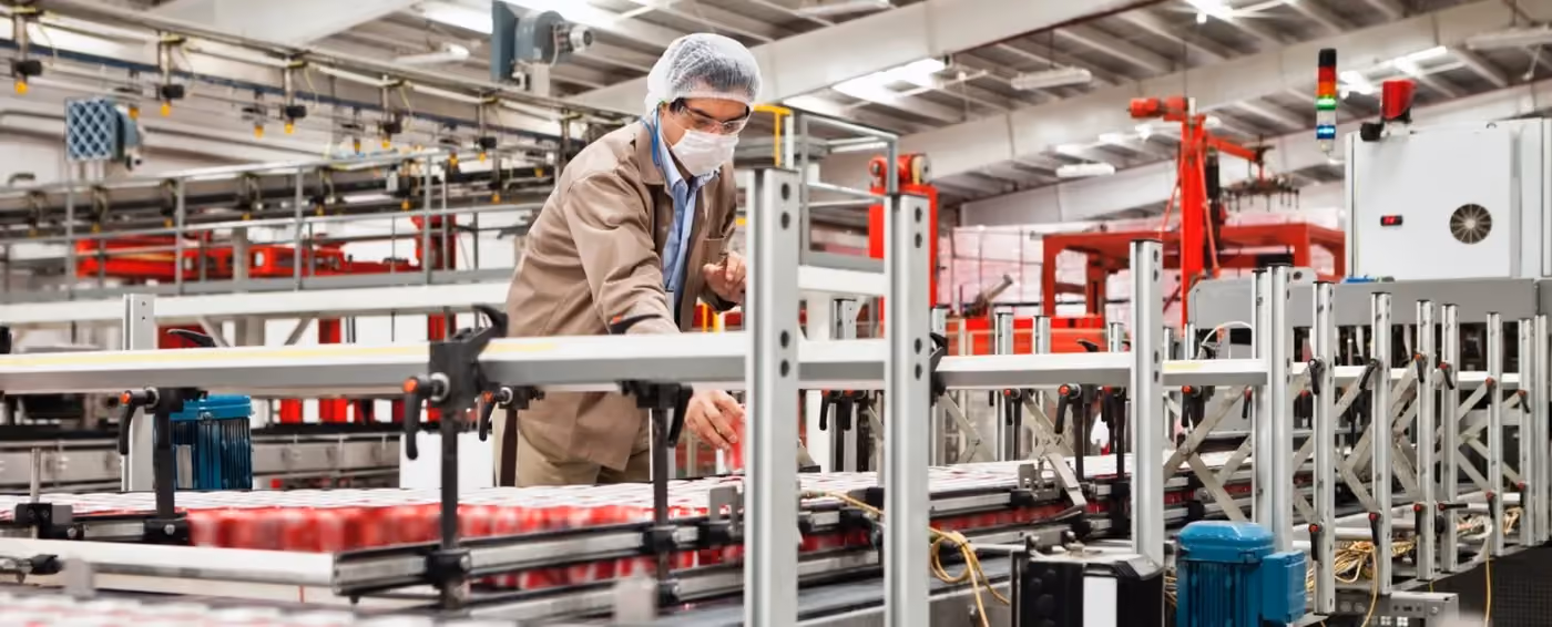 Worker wearing safety gear inspects cans on a conveyor belt in a large industrial factory.