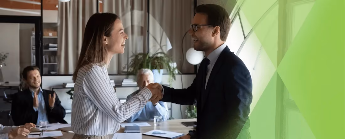 A man and woman shaking hands in an office while colleagues applaud around a meeting table.
