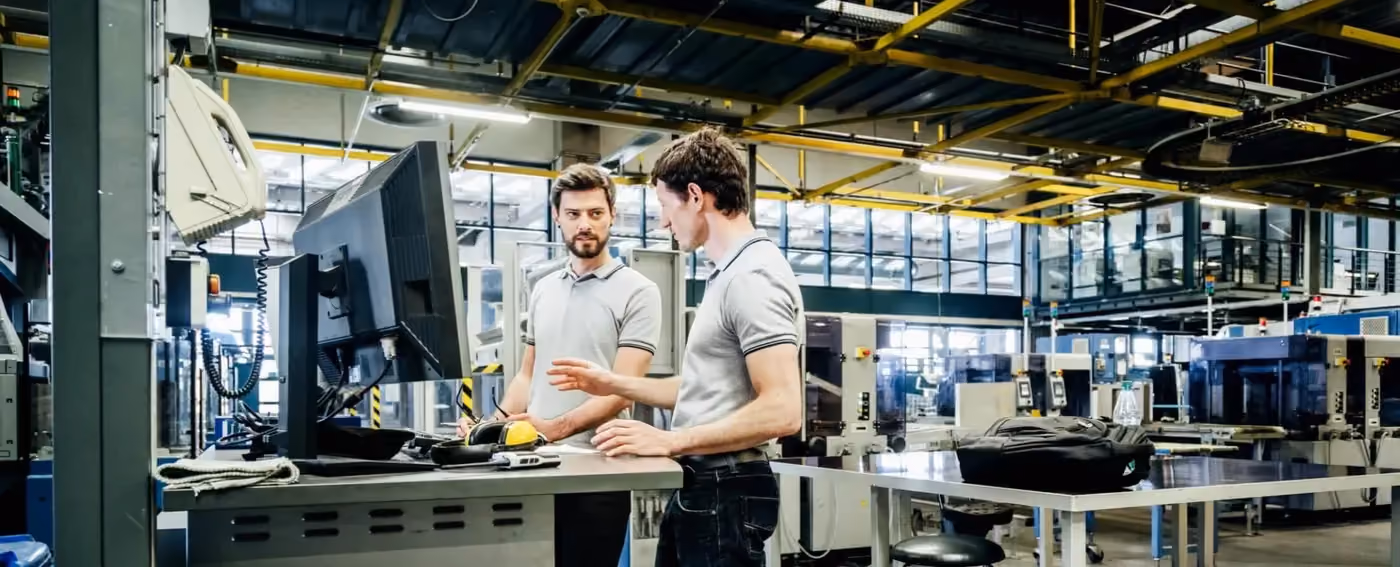 Two men in a factory discuss while looking at a large computer monitor on a workstation.