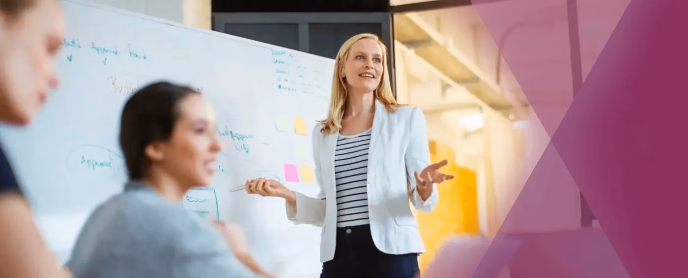 Woman in white blazer leading a presentation with a marker and whiteboard with notes behind her.