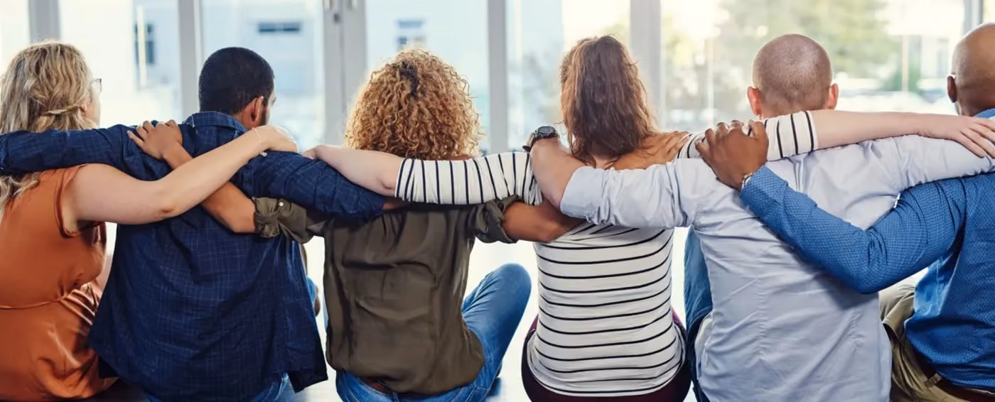 Six diverse people sitting side by side with arms around each other’s shoulders in a supportive group.