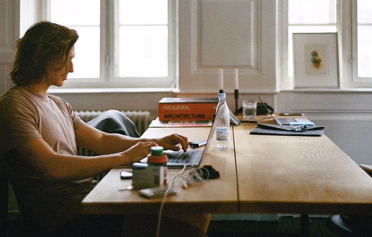 Person typing on laptop at a wooden desk with books, water bottle, and other items near a window.