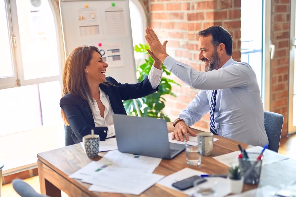 Two colleagues smiling and giving a high five across a desk with documents and a laptop.