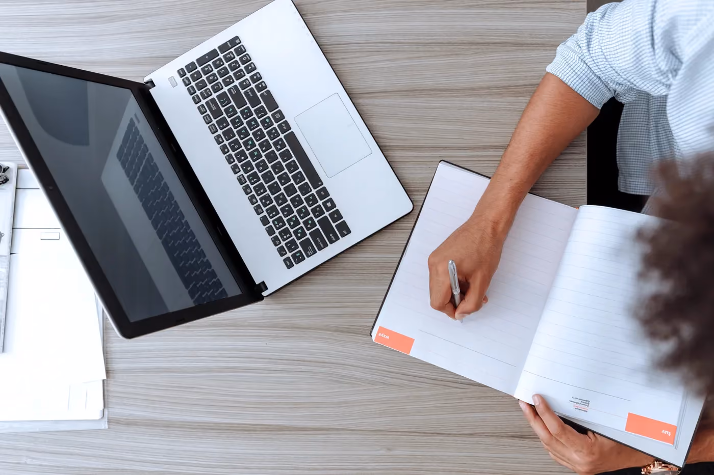 Person writing in an open notebook on a wood desk next to a laptop and papers.