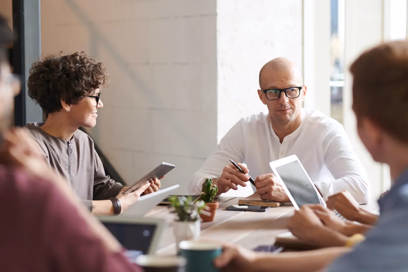 A diverse group in a meeting around a table using tablets, notebooks, and coffee cups.