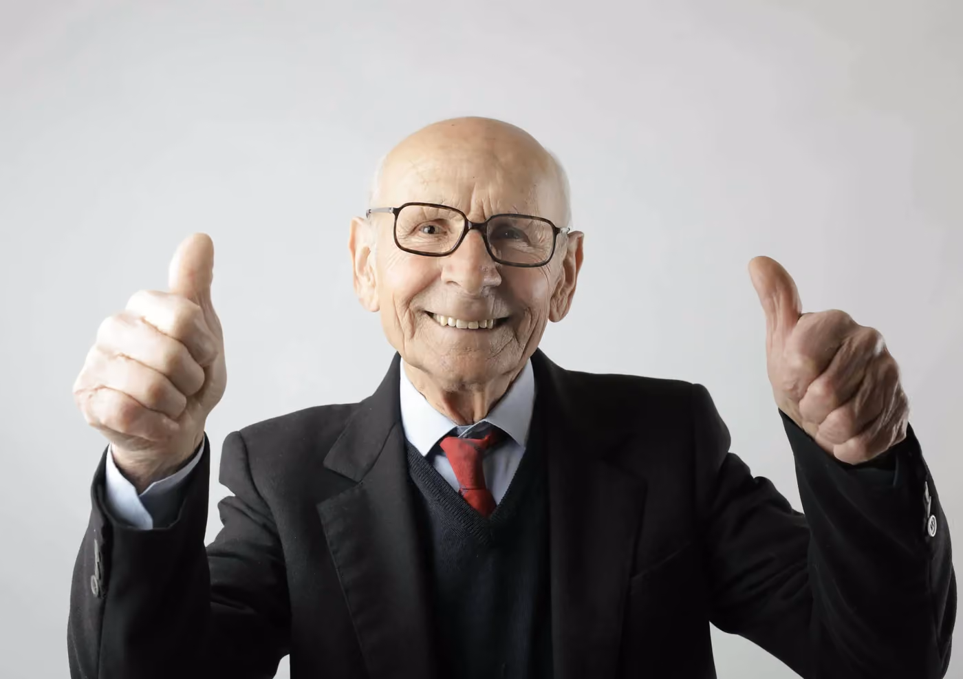 Smiling elderly man in suit and glasses showing thumbs up with both hands.
