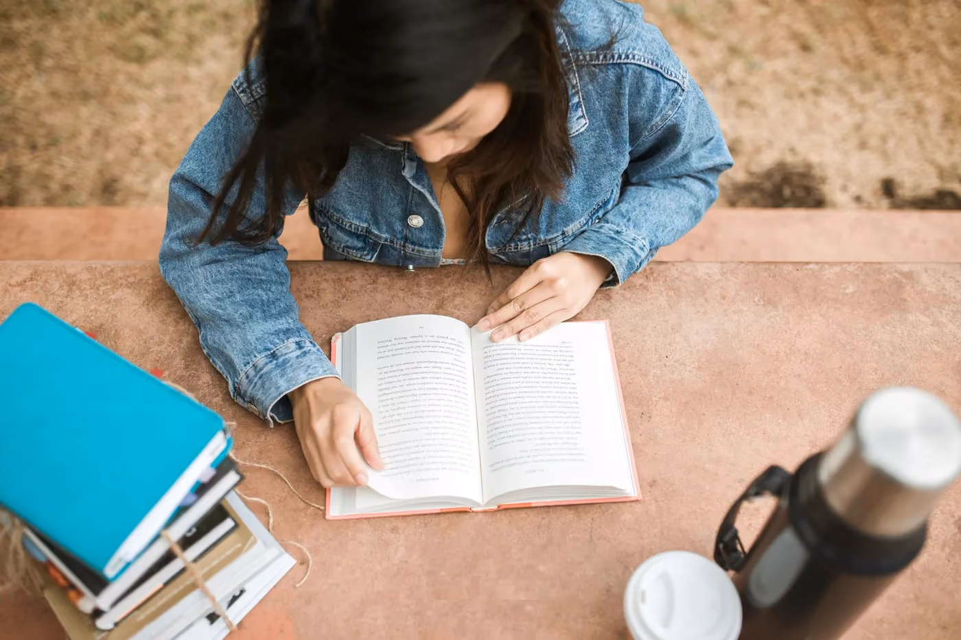 Person reading an open book at a table with a stack of books and a thermos nearby.