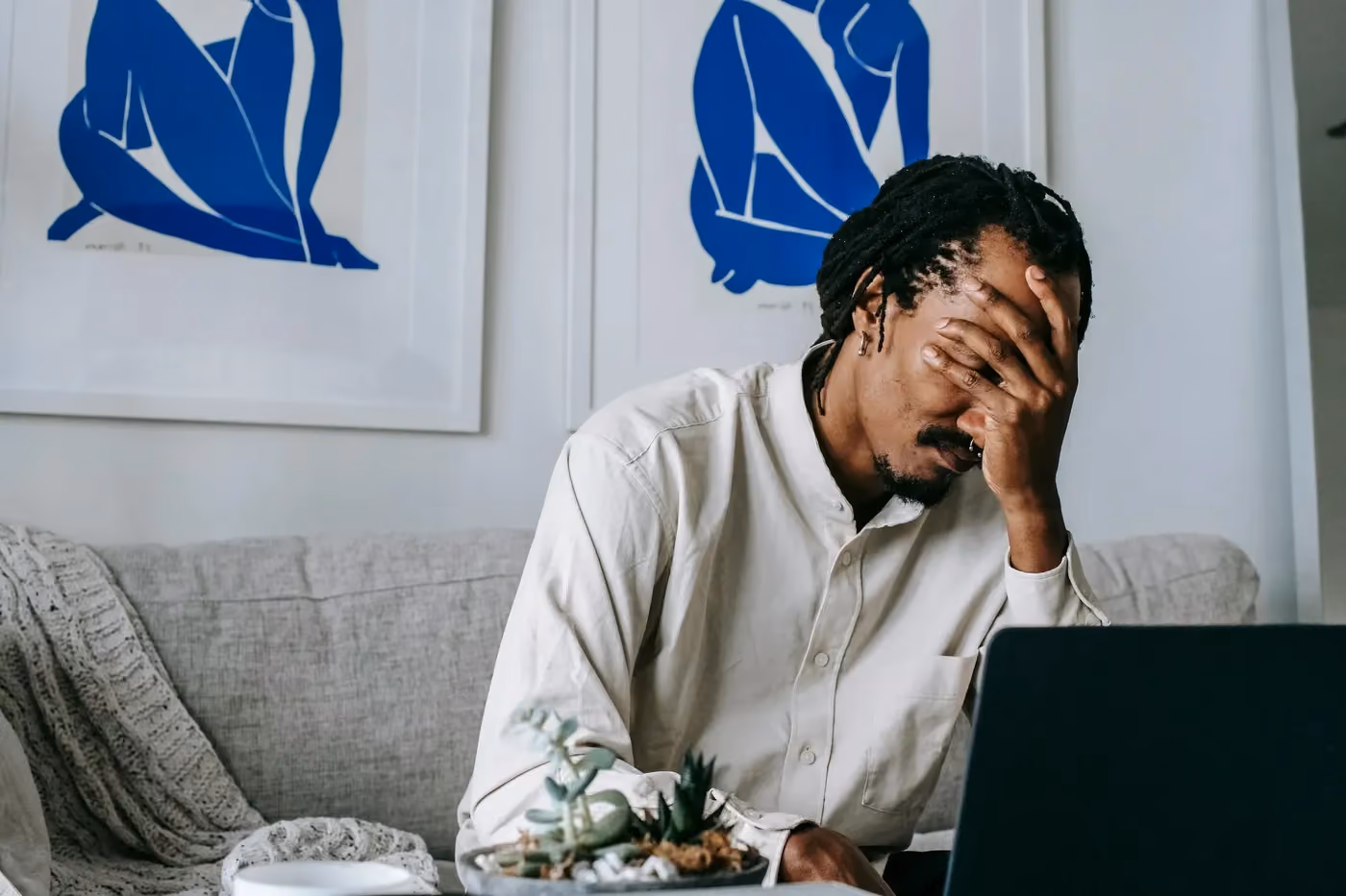 Man sitting on couch with hand covering his face in front of a laptop, appearing stressed.