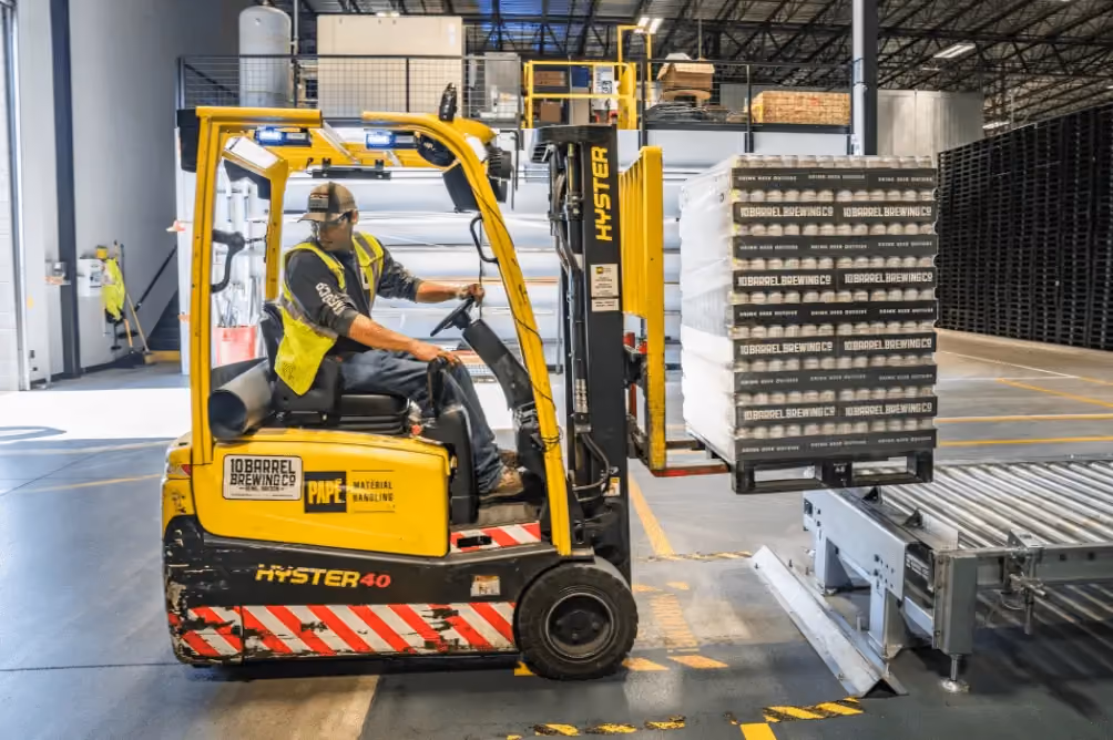 Worker driving a yellow forklift carrying pallets labeled 10 Barrel Brewing Co in warehouse.