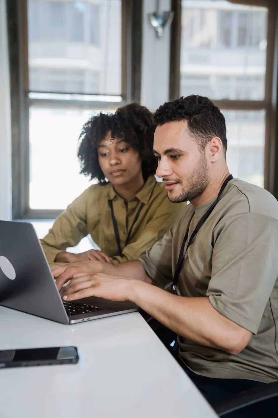 Two coworkers at a table collaborating on a laptop in an office with large windows.