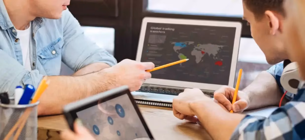 Two people collaborating at a desk with laptops showing global trading maps and charts.