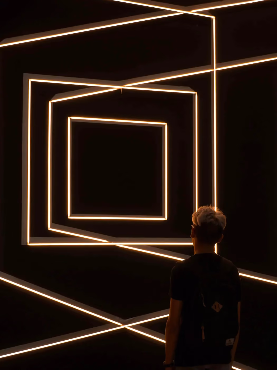 Person with backpack looking at illuminated geometric square light installation on black walls.