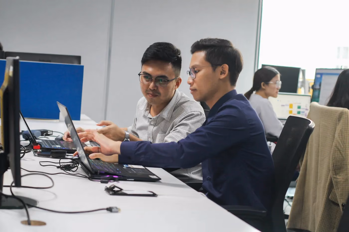 Two men collaborate on laptops in a modern office with colleagues working in the background.