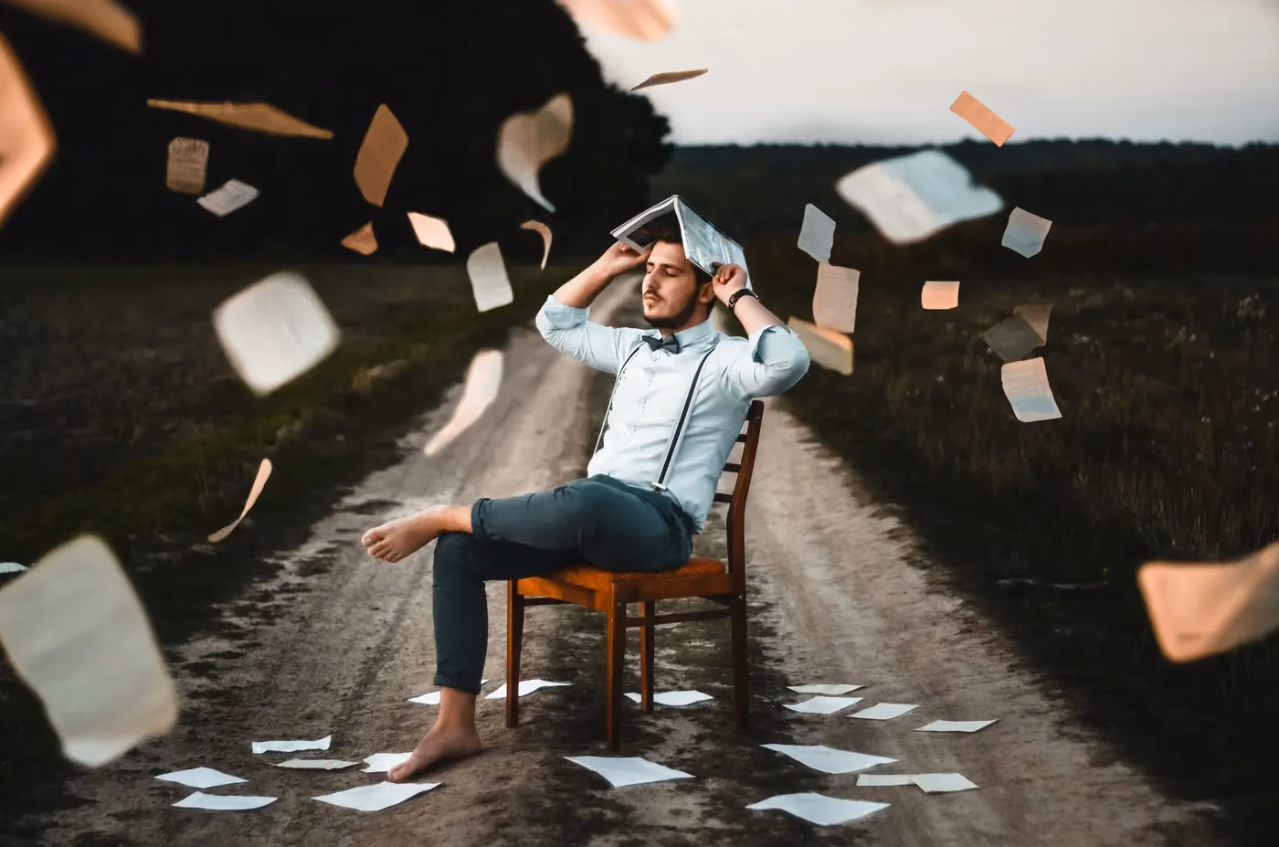 A barefoot man sits on a chair on a dirt road holding a book over his head as papers fly around him.