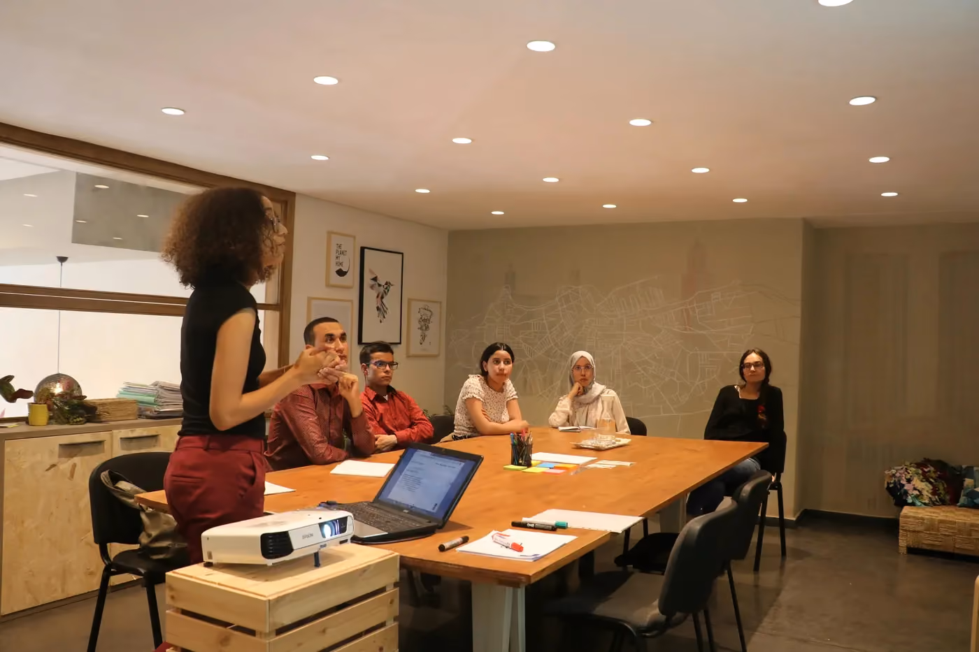 Woman standing and speaking to a group of five seated people around a conference table with a laptop and projector.