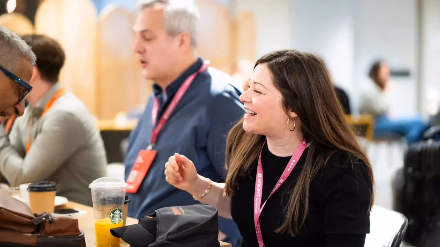 Smiling woman with dark hair and pink lanyard conversing with seated men at a table with drinks.