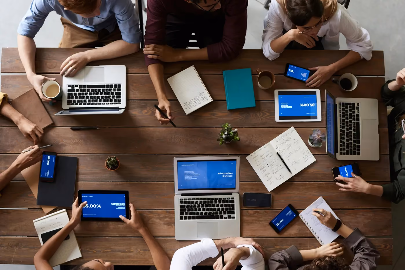 Top view of people working at a wood table with laptops, tablets, smartphones, notebooks, and coffee mugs.