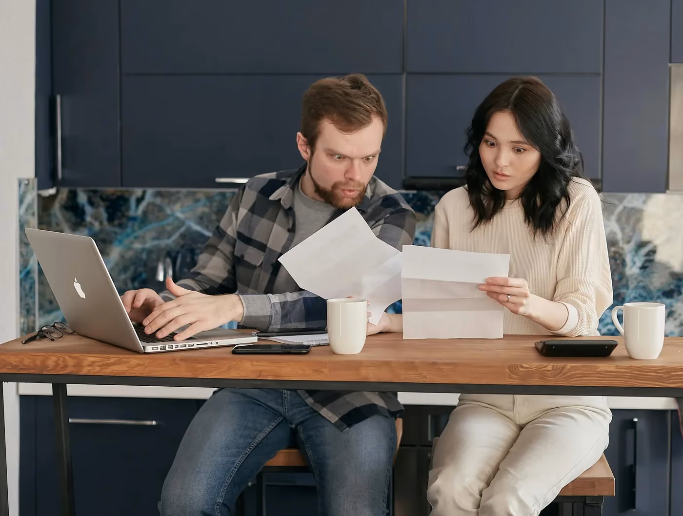 Man and woman seated at a table looking surprised while reviewing papers with a laptop and coffee mugs.