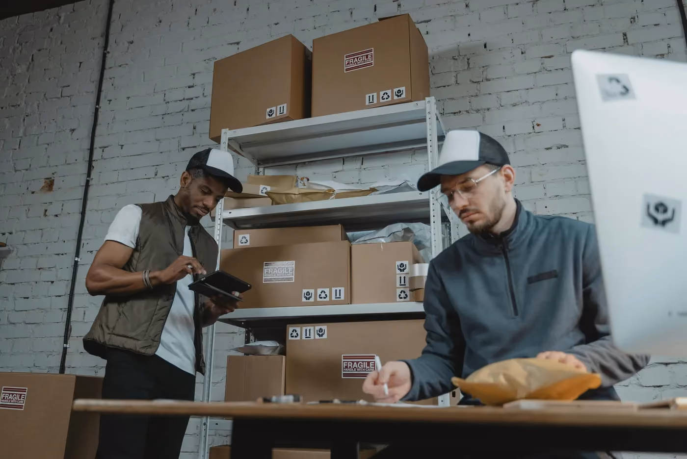 Two men working in a warehouse with cardboard boxes, one using a tablet, the other handling a package.