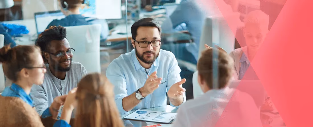 Five diverse coworkers seated at a table in discussion with charts and graphs visible on paper.