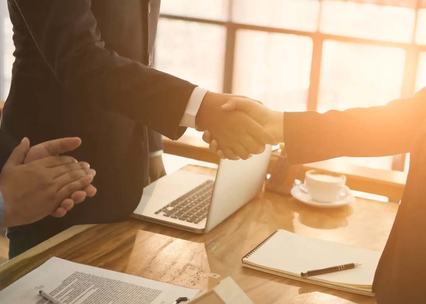 Two people in suits shaking hands over a desk with laptop, papers, and coffee cup in sunlight.