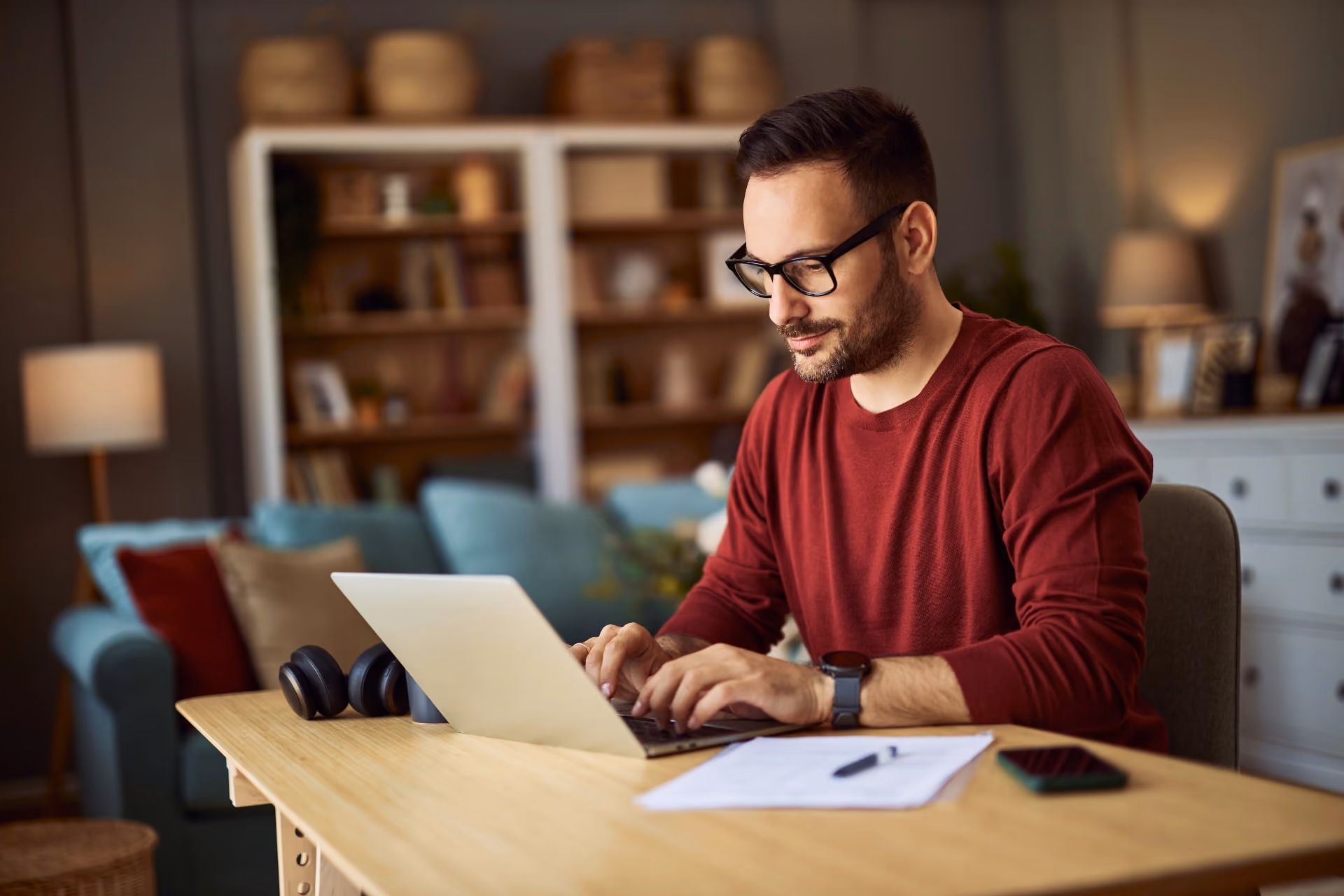 Man in red sweater wearing glasses working on laptop at home desk with papers and phone nearby.