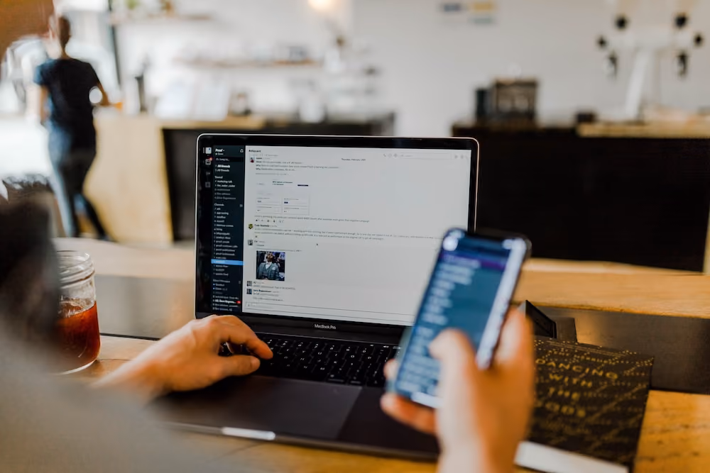 Person using MacBook Pro with messaging app on screen and holding smartphone at wooden table.