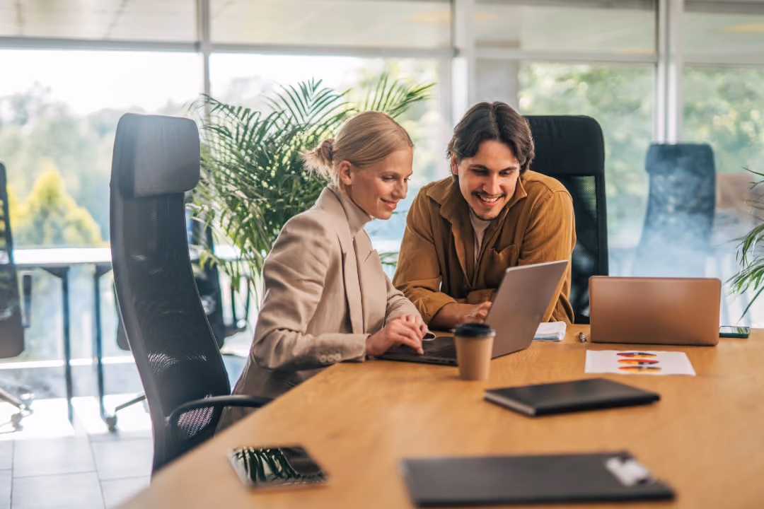 Man and woman collaborate happily on laptop at office table with coffee and documents nearby.