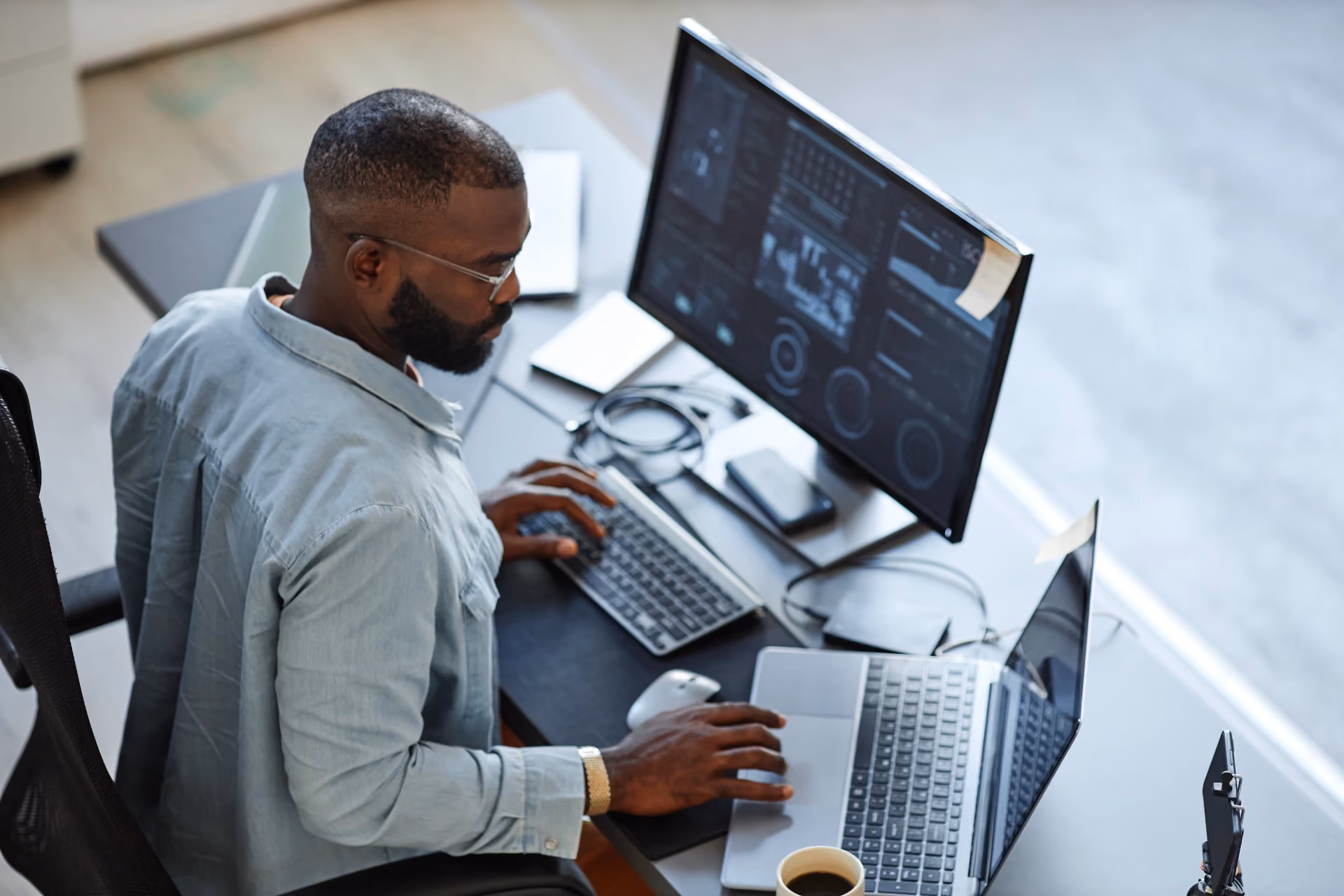Man with glasses working on a laptop and desktop computer displaying data charts.