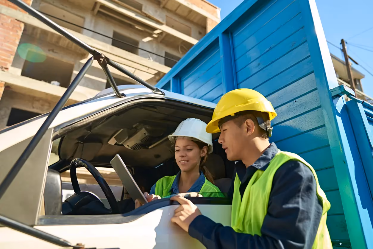 Two construction workers in hard hats and vests using a tablet in a truck at a building site.