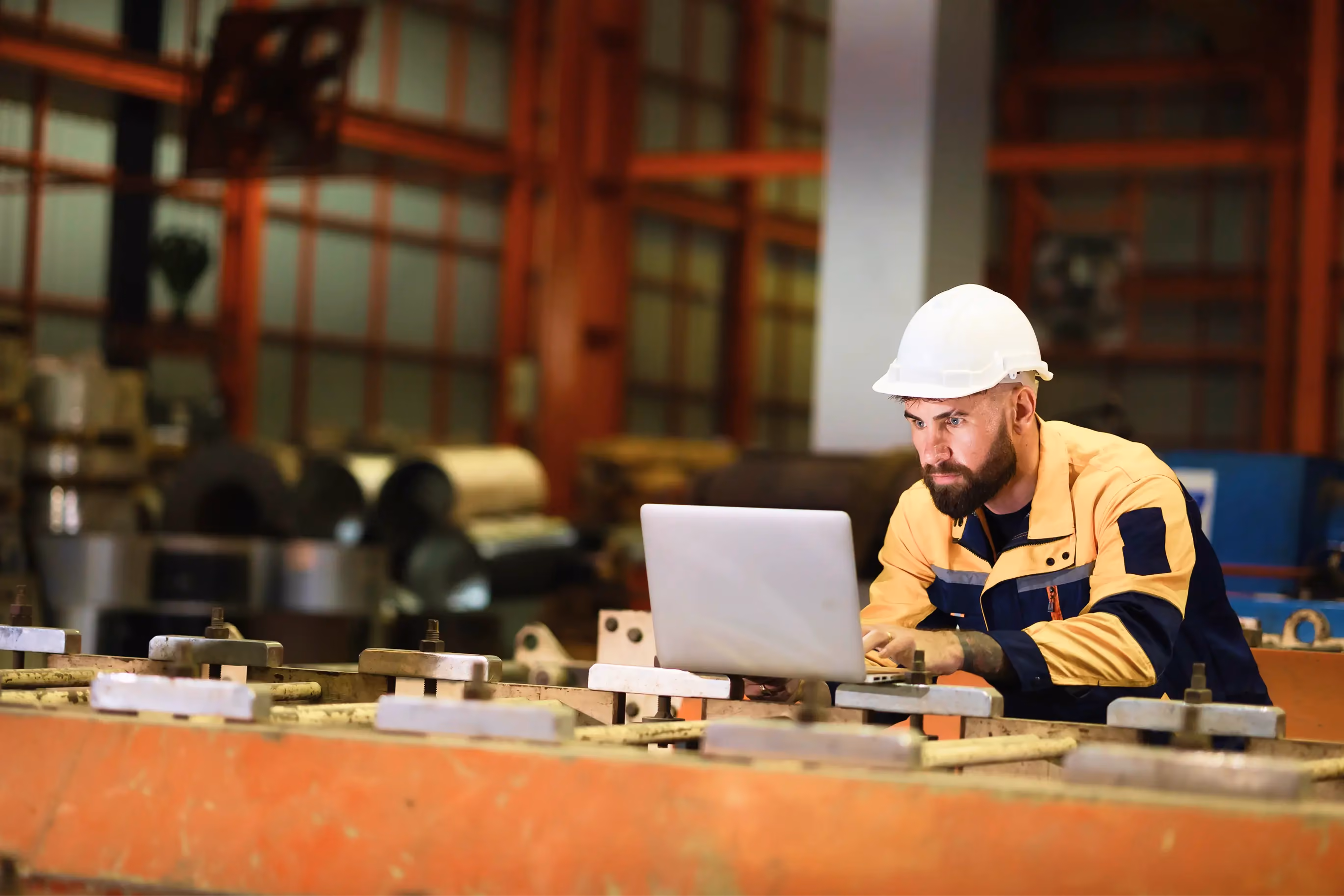 Male factory worker in a hard hat using a laptop in an industrial setting with metal coils in the background.