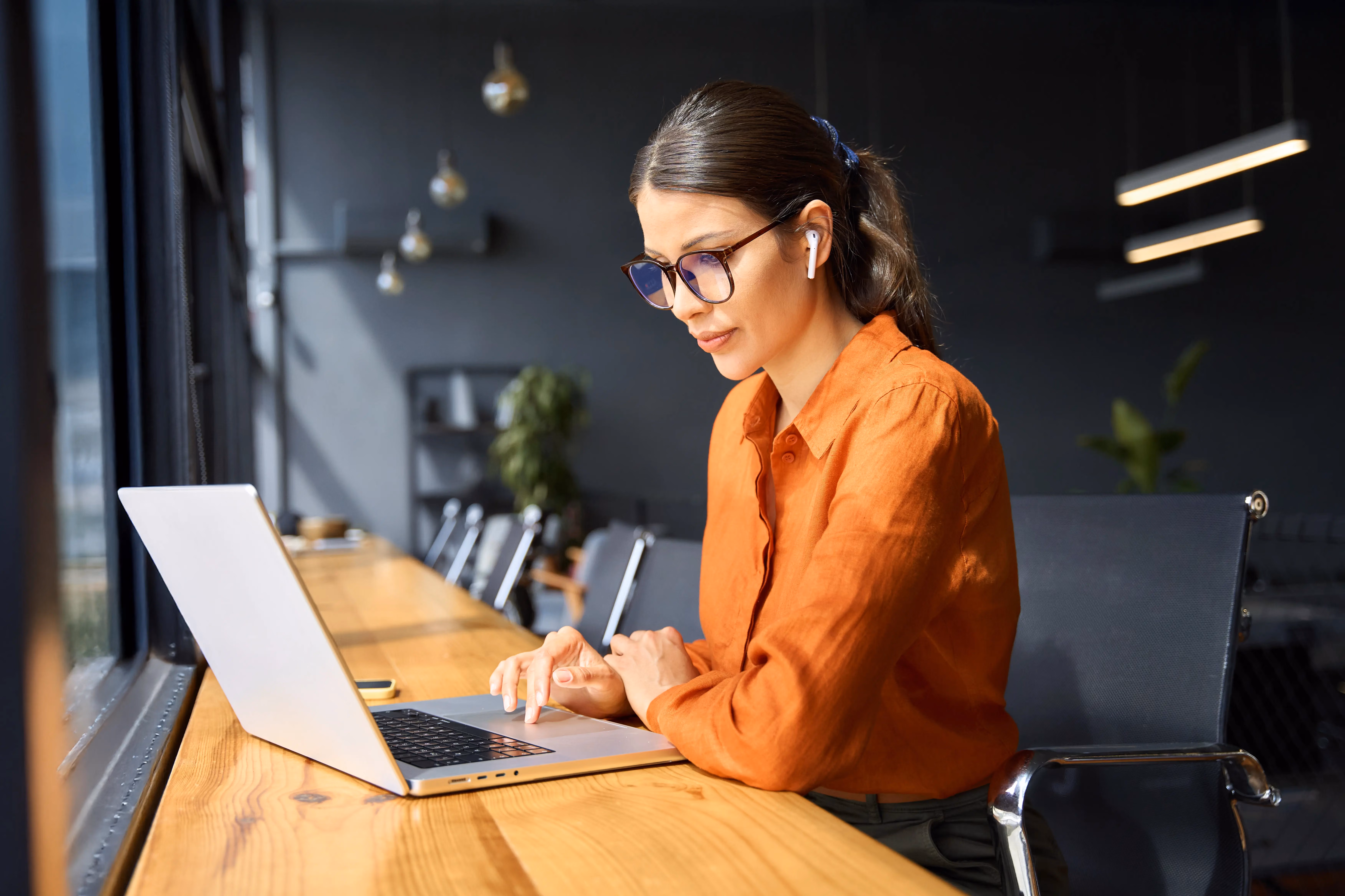 Woman wearing glasses and orange shirt working on a laptop at a wooden desk by a window.