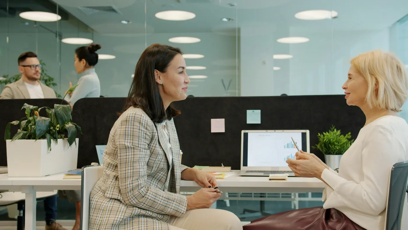 Two colleagues in a one-on-one conversation at a laptop in a bright open-plan office, one sharing feedback while the other listens.