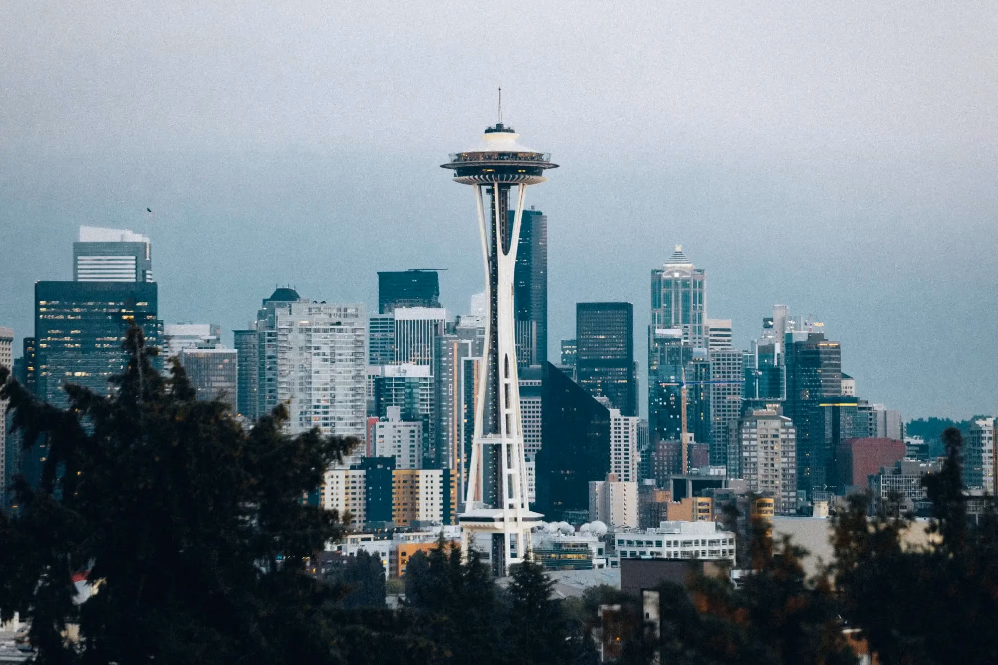 Seattle city skyline featuring the Space Needle prominently in the center, surrounded by various modern high-rise buildings, seen against a cloudy sky with trees in the foreground.