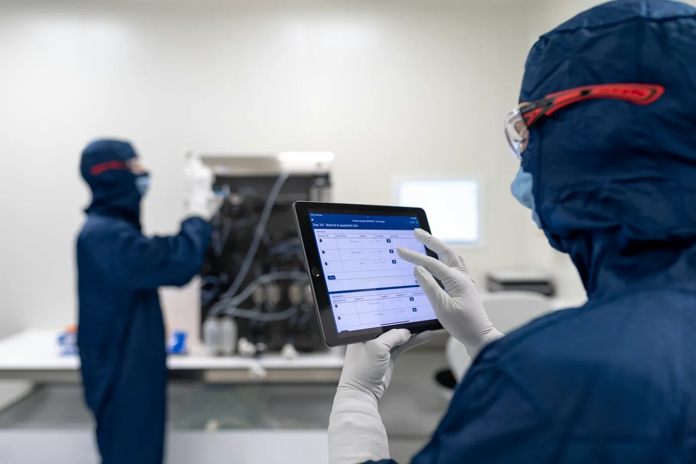 A lab technician in a cleanroom suit and gloves follows a step-by-step procedure on a tablet while a colleague works at equipment in the background.