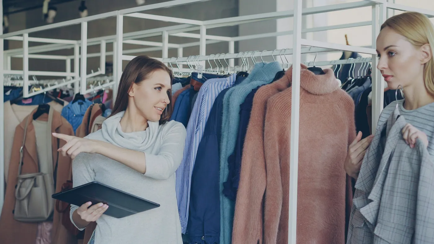 A clothing store employee holding a tablet points toward a clothing rack while talking with a colleague who is holding a garment.